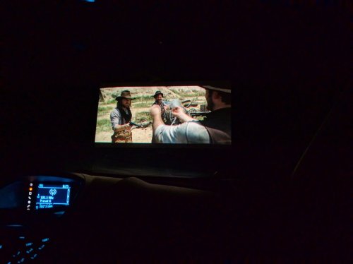 A dark scene showing a movie playing on a screen, featuring two characters in a standoff in a desert setting.
