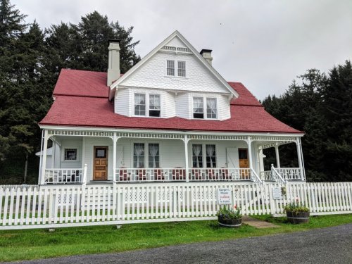 A large white house with a red roof, surrounded by a white picket fence and trees in the background.