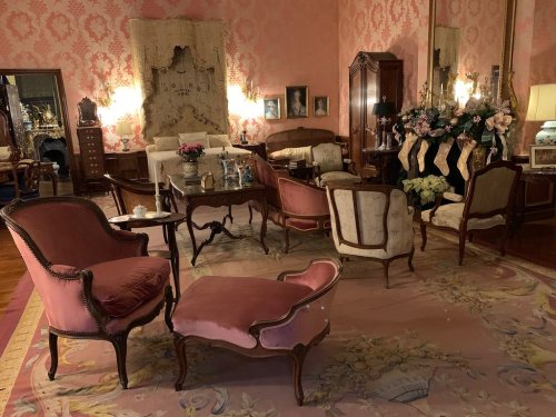 A vintage living room with pink wallpaper, elegant furniture, and floral decorations, creating a cozy, classic atmosphere.