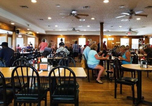 A busy restaurant interior with diners at tables, wooden walls, and ceiling fans, creating a lively atmosphere.
