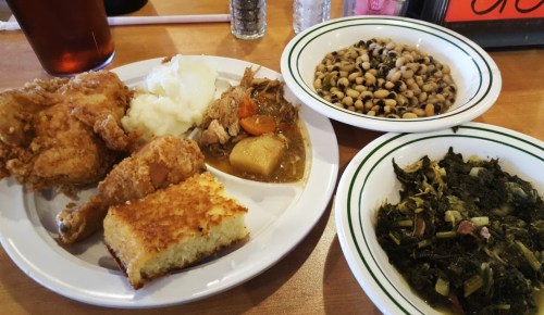 A plate of Southern comfort food featuring fried chicken, mashed potatoes, cornbread, black-eyed peas, and collard greens.