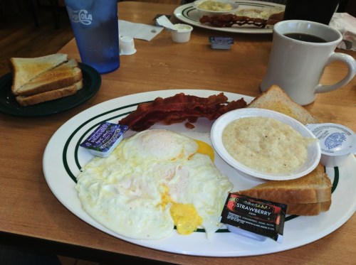 A breakfast plate with eggs, bacon, grits, toast, and condiments, alongside a cup of coffee and a blue water glass.