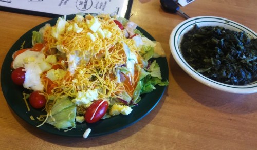 A colorful salad topped with cheese, eggs, and tomatoes, served with a side of collard greens on a wooden table.