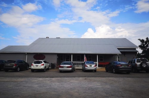A gray building with a metal roof and several parked cars in front, under a blue sky with clouds.