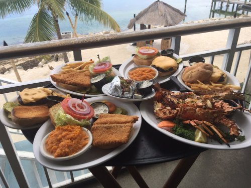 A variety of seafood and burgers served on plates, with sides of fries, salad, and sweet potato, against a waterfront backdrop.