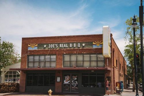 A brick building with a sign reading "Joe's Real B-B-Q" under a clear blue sky, featuring a vintage style and nearby trees.