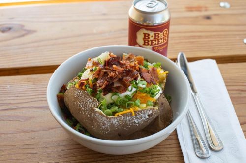 A loaded baked potato topped with cheese, green onions, bacon, and a can of beer on a wooden table.