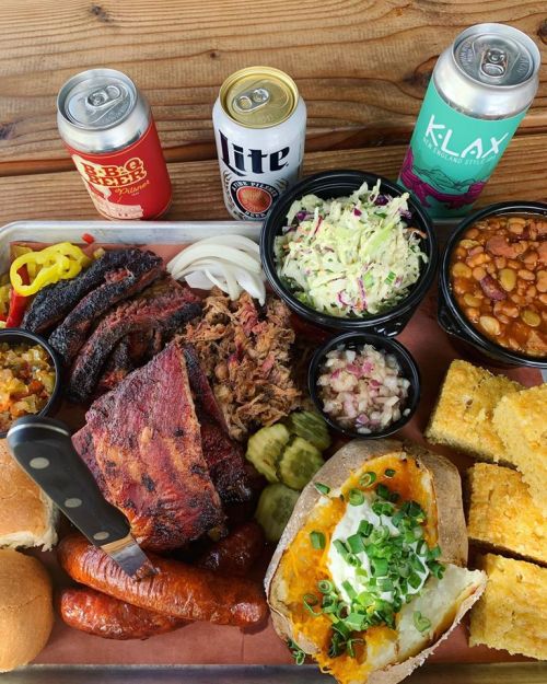 A platter of barbecue meats, sides like coleslaw and baked beans, with drinks and cornbread on a wooden table.