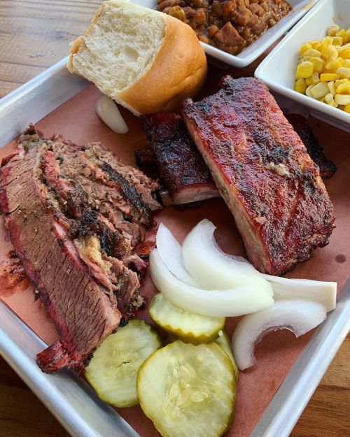 A tray of barbecue featuring brisket, ribs, pickles, onions, and a bun, served with sides of baked beans and corn.