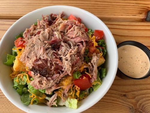 A bowl of salad topped with shredded meat, tomatoes, cheese, and a side of dressing on a wooden table.