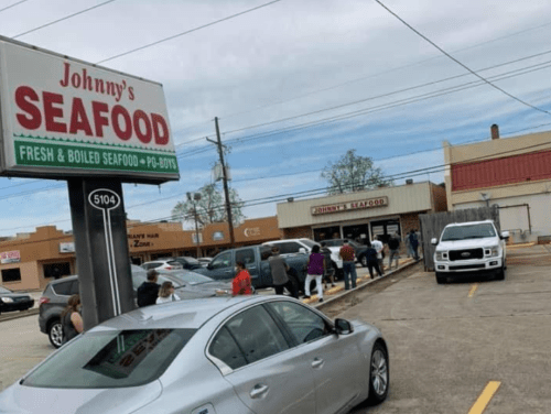 A busy street scene featuring a seafood restaurant with a long line of customers outside and parked cars nearby.