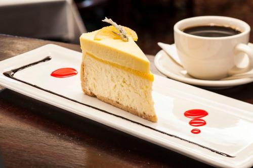 A slice of lemon cheesecake on a white plate, accompanied by a cup of coffee. Red sauce drizzled on the plate.