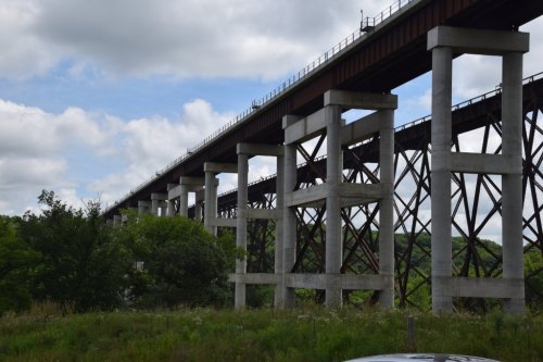 A tall railway bridge supported by concrete and steel pillars, set against a cloudy sky and green landscape.
