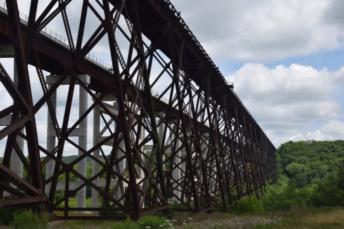 A large, rusted railway trestle bridge spans over green foliage under a cloudy sky.