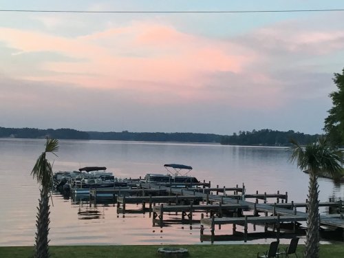 A serene lake at sunset, with boats docked at a wooden pier and palm trees in the foreground.