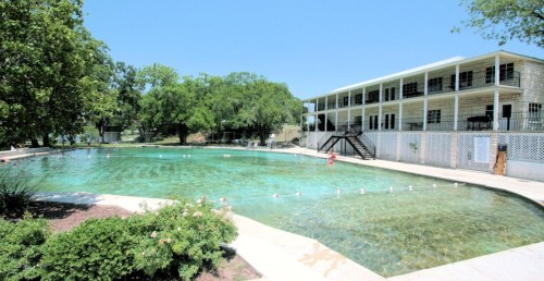 A clear swimming pool surrounded by greenery and a white building with a balcony in the background.