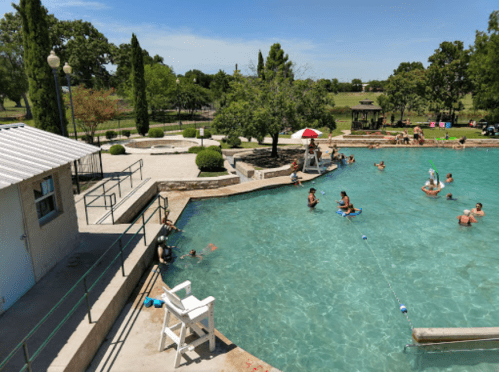 A sunny pool scene with people swimming, lounging, and enjoying the water, surrounded by trees and grassy areas.