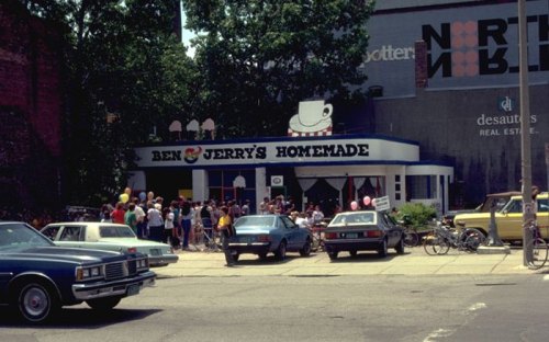 A busy Ben & Jerry's ice cream shop with a crowd outside, vintage cars parked nearby, and trees in the background.