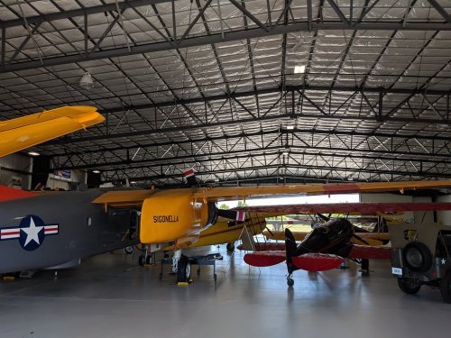 A spacious hangar with vintage aircraft, including a yellow biplane and a black plane, under a metal roof.