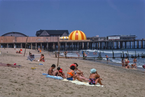 A sunny beach scene with people sunbathing, a pier in the background, and colorful beach umbrellas.