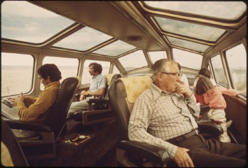 Passengers relax in a train's observation car, enjoying views through large windows on a scenic journey.