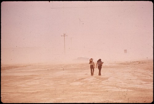 Two figures walk through a dusty landscape, with low visibility and power lines in the background.