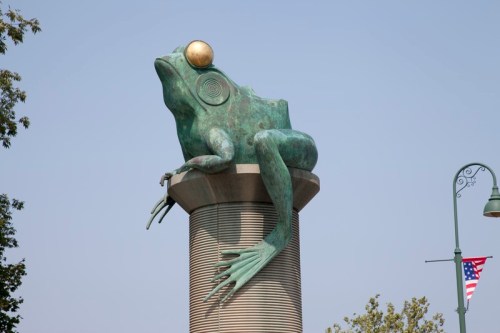 A large bronze frog sculpture perched on a cylindrical pedestal, with a golden orb on its head against a clear sky.