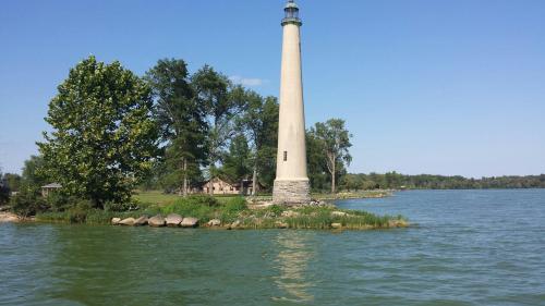 A tall lighthouse stands on a green island surrounded by calm water and trees under a clear blue sky.