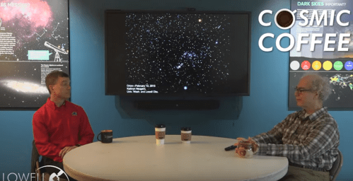 Two people sit at a table with coffee cups, discussing astronomy in front of a starry sky presentation on a screen.