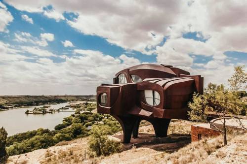 A unique, modern house with a sculptural design, perched on a hillside overlooking a river and lush greenery.