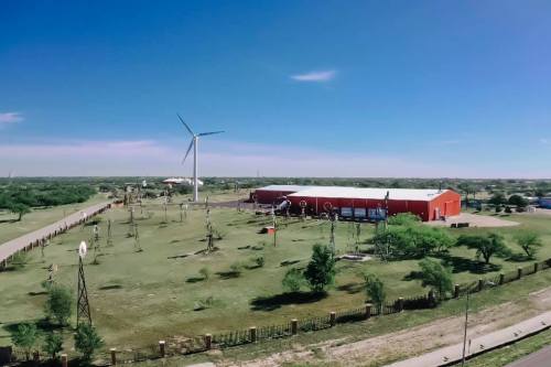 Aerial view of a red building surrounded by green fields, wind turbines, and vintage windmills under a clear blue sky.