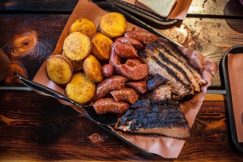 A platter of grilled meats including brisket and sausages, served with golden cornbread on a wooden table.