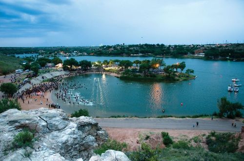 Aerial view of a lake with a crowd gathered on the shore, surrounded by trees and boats on the water under a cloudy sky.