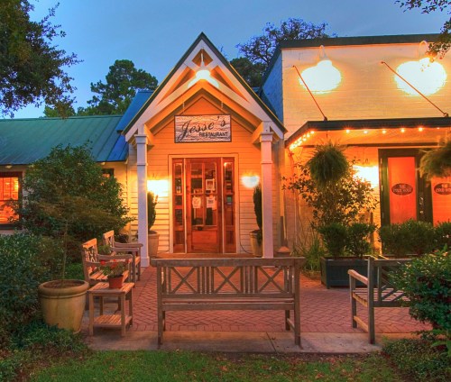 A charming restaurant exterior with warm lighting, benches, and greenery, inviting guests to enter.