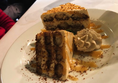 A slice of layered dessert with cookies, cream, and chocolate shavings, drizzled with caramel sauce and topped with whipped cream.