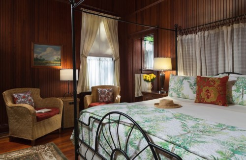 Cozy bedroom with wooden walls, a four-poster bed, tropical decor, and a seating area with wicker chairs.