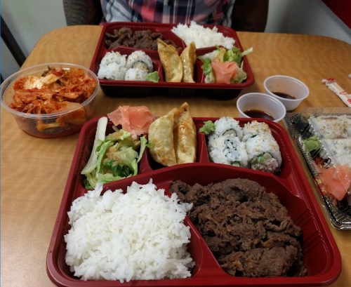 Two red trays of Asian cuisine featuring rice, beef, sushi, dumplings, salad, and kimchi on a wooden table.