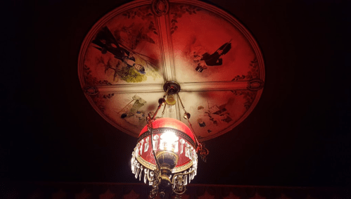 A decorative ceiling with vintage paintings and a red chandelier adorned with crystals, illuminated softly.