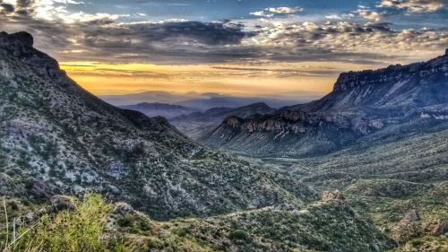 A scenic view of rolling hills and valleys under a colorful sunset sky, with distant mountains in the background.
