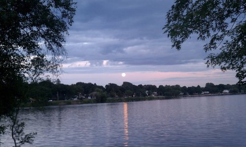 A serene lake scene at dusk, with a full moon reflecting on the water and clouds in the sky.