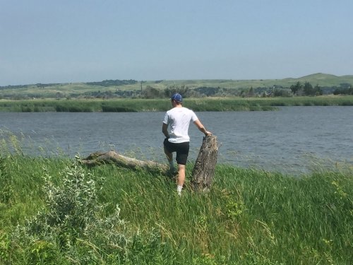 A person stands on a log by a river, surrounded by tall grass and hills in the background under a clear sky.