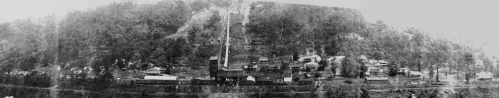 A historic black-and-white panorama of a mining town nestled in a mountainous landscape, featuring buildings and rail tracks.
