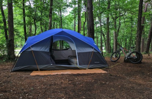 A blue and gray tent set up in a forest, with a bike parked nearby on a brown mat.