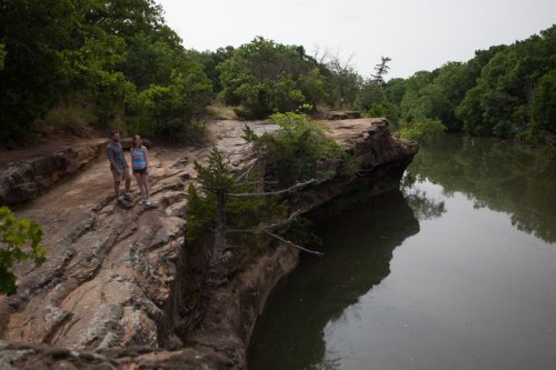 Two people stand on a rocky ledge by a calm river, surrounded by lush green trees and a serene landscape.