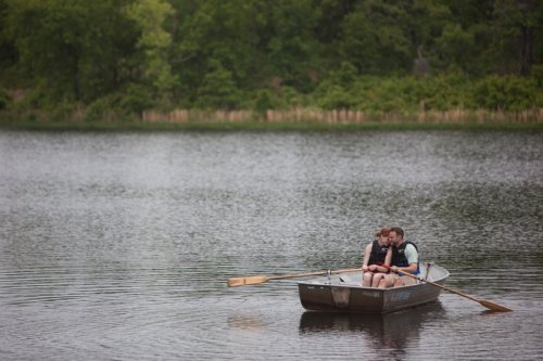 A couple sits in a small boat on a calm lake, surrounded by lush greenery, enjoying a peaceful moment together.