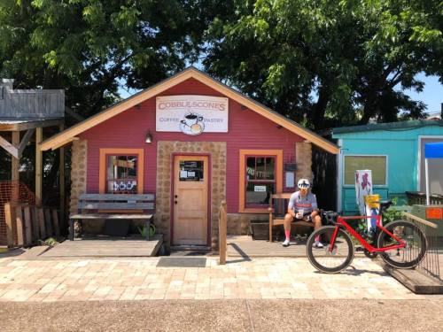 A quaint coffee shop named "Cobble Scones" with a cyclist resting outside and a red bike parked nearby.