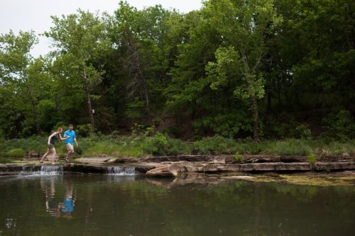 Two people playfully jump across rocks by a calm river, surrounded by lush green trees and foliage.