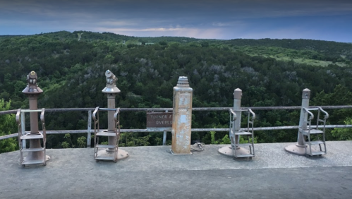 View from an overlook with binoculars mounted on a railing, surrounded by lush green hills and trees.