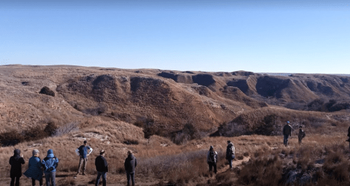 A group of people stands on a hillside overlooking a rugged, grassy landscape with canyons under a clear blue sky.