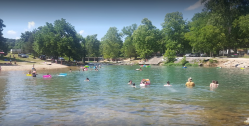 People swimming and relaxing in a clear river surrounded by trees, with colorful floats and a sandy shore.